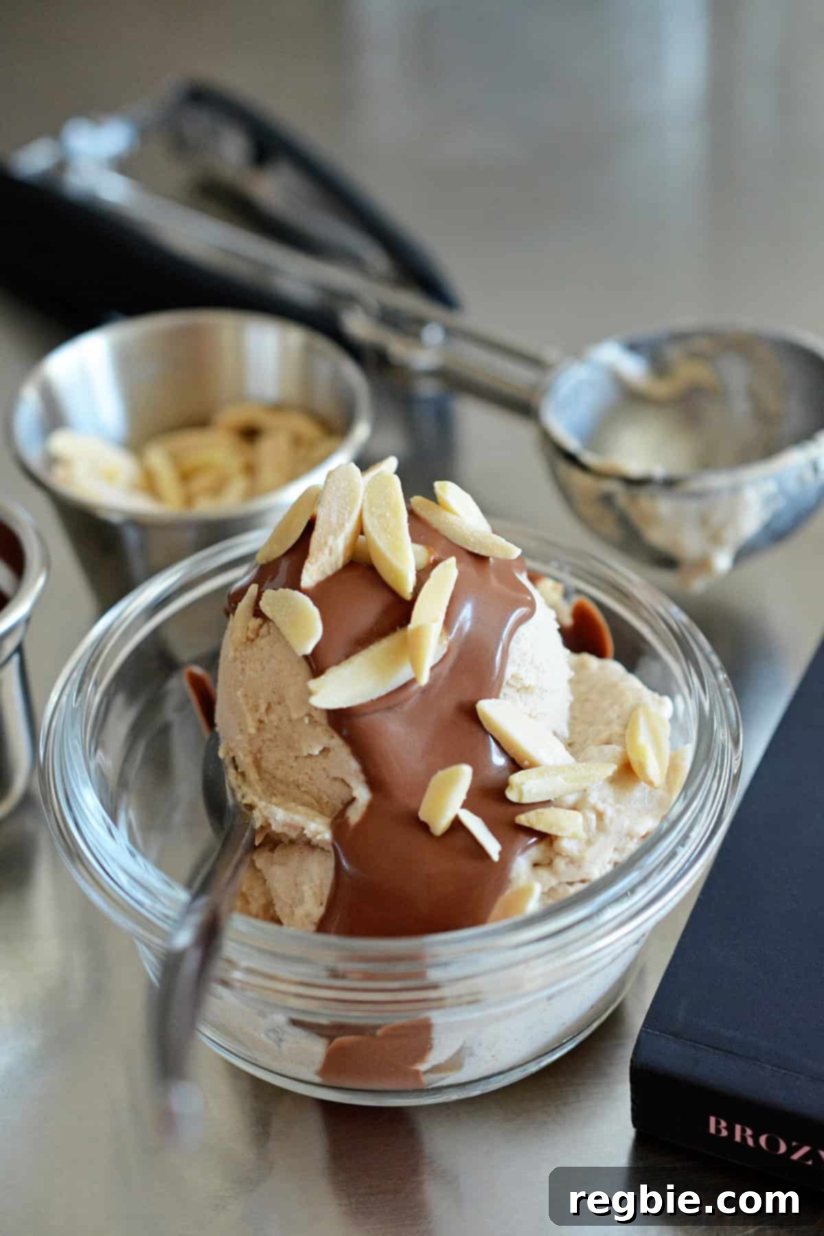 A side view of a scoop of homemade dairy-free vanilla ice cream topped with luxurious chocolate ganache and toasted almond slivers, surrounded by fresh ingredients.