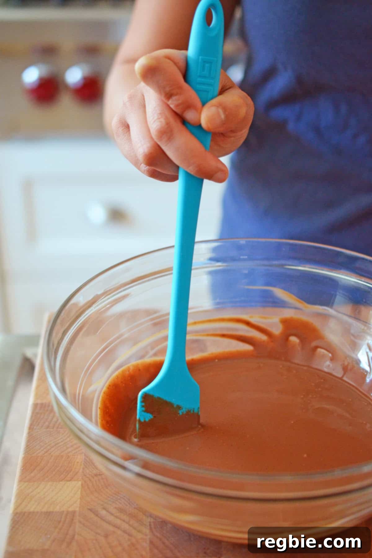 A blue spatula stirring rich chocolate ganache in a glass mixing bowl, set on a wooden cutting board, creating a smooth and glossy texture.
