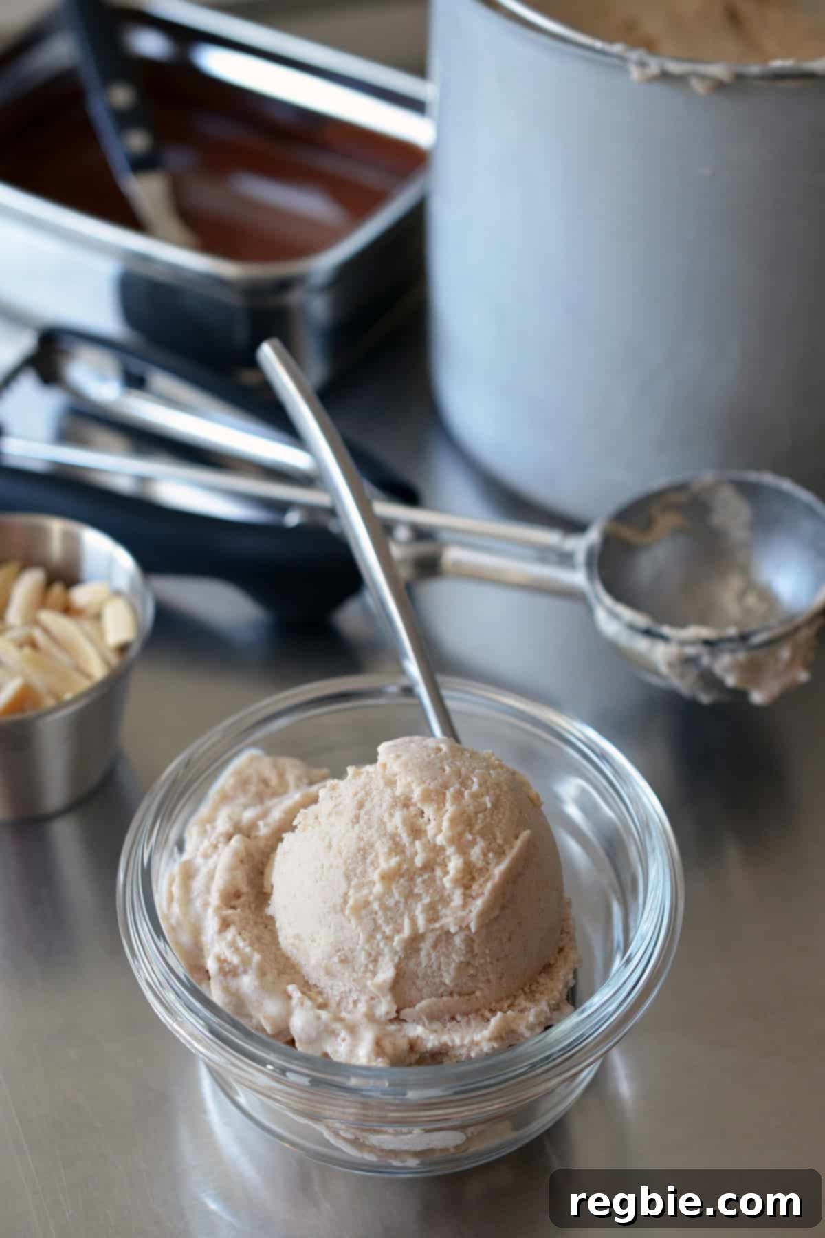 A scoop of vanilla paleo ice cream in a small glass bowl, positioned next to an ice cream scooper, a container of chocolate sauce, and toasted almond slivers.