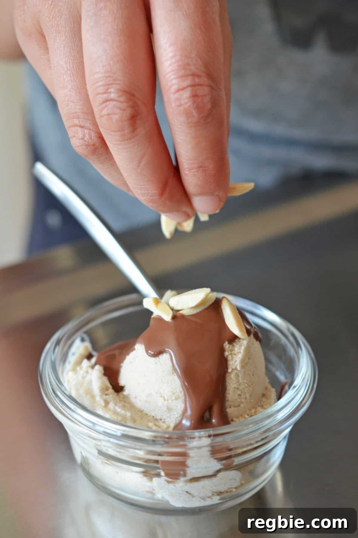 A hand gracefully sprinkling toasted almond slivers over a scoop of homemade paleo ice cream, already topped with rich chocolate sauce.
