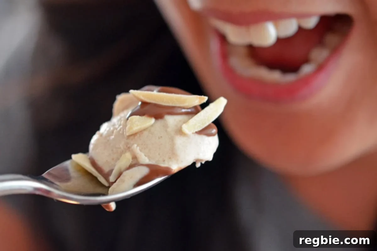 A spoonful of homemade vanilla ice cream topped with chocolate ganache and almond slivers approaches an open mouth, ready for a delicious bite.