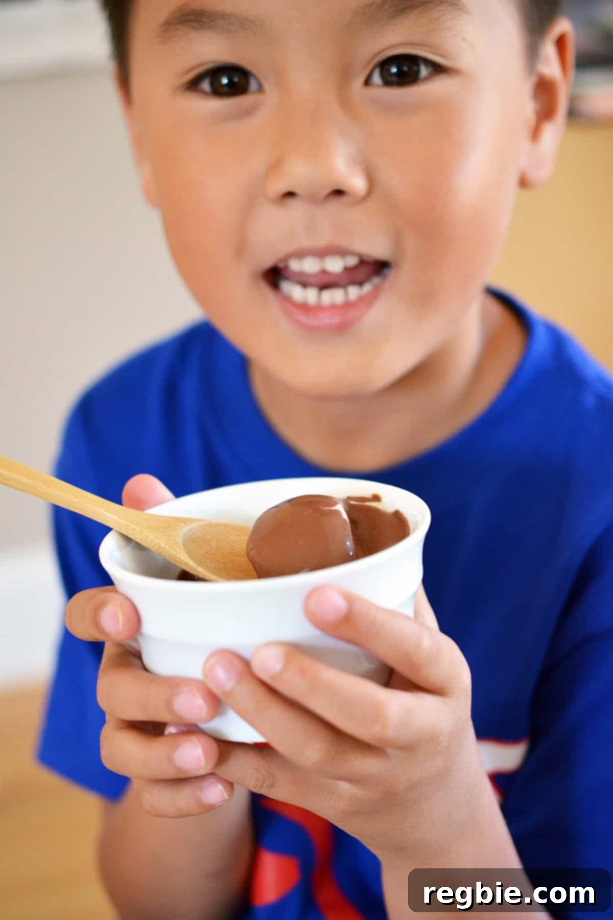 A smiling Asian boy holding a white bowl brimming with dairy-free vanilla ice cream, generously topped with chocolate ganache, looking utterly delighted.