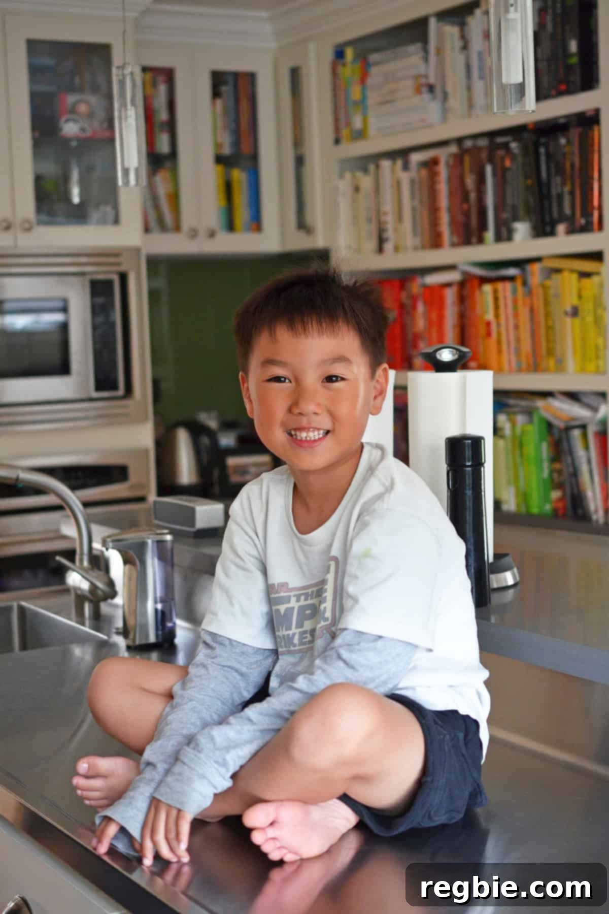 A smiling Asian boy sitting cross-legged on a kitchen counter, full of joy and anticipation for homemade ice cream.