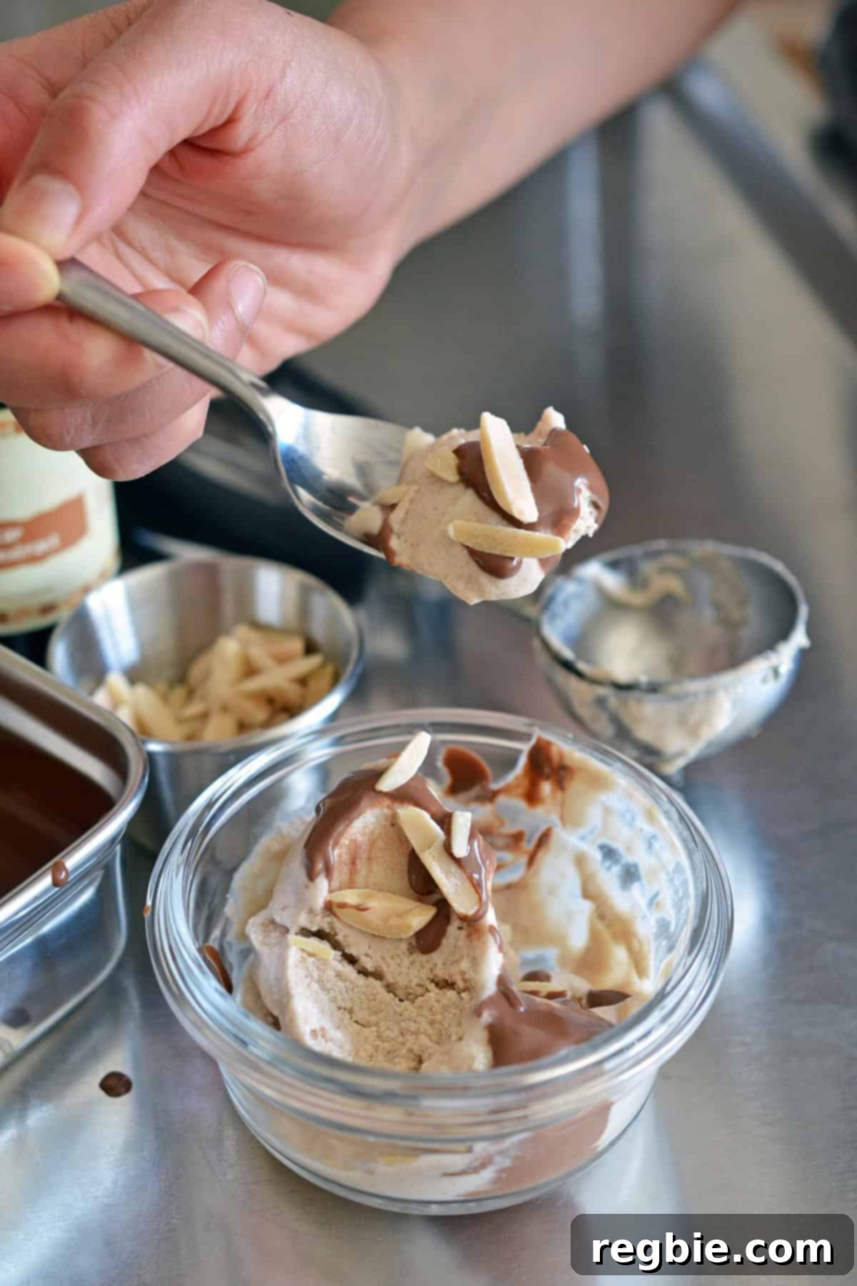 A hand taking a spoonful from a delightful scoop of homemade dairy-free vanilla ice cream, generously topped with chocolate ganache and crisp almond slivers.