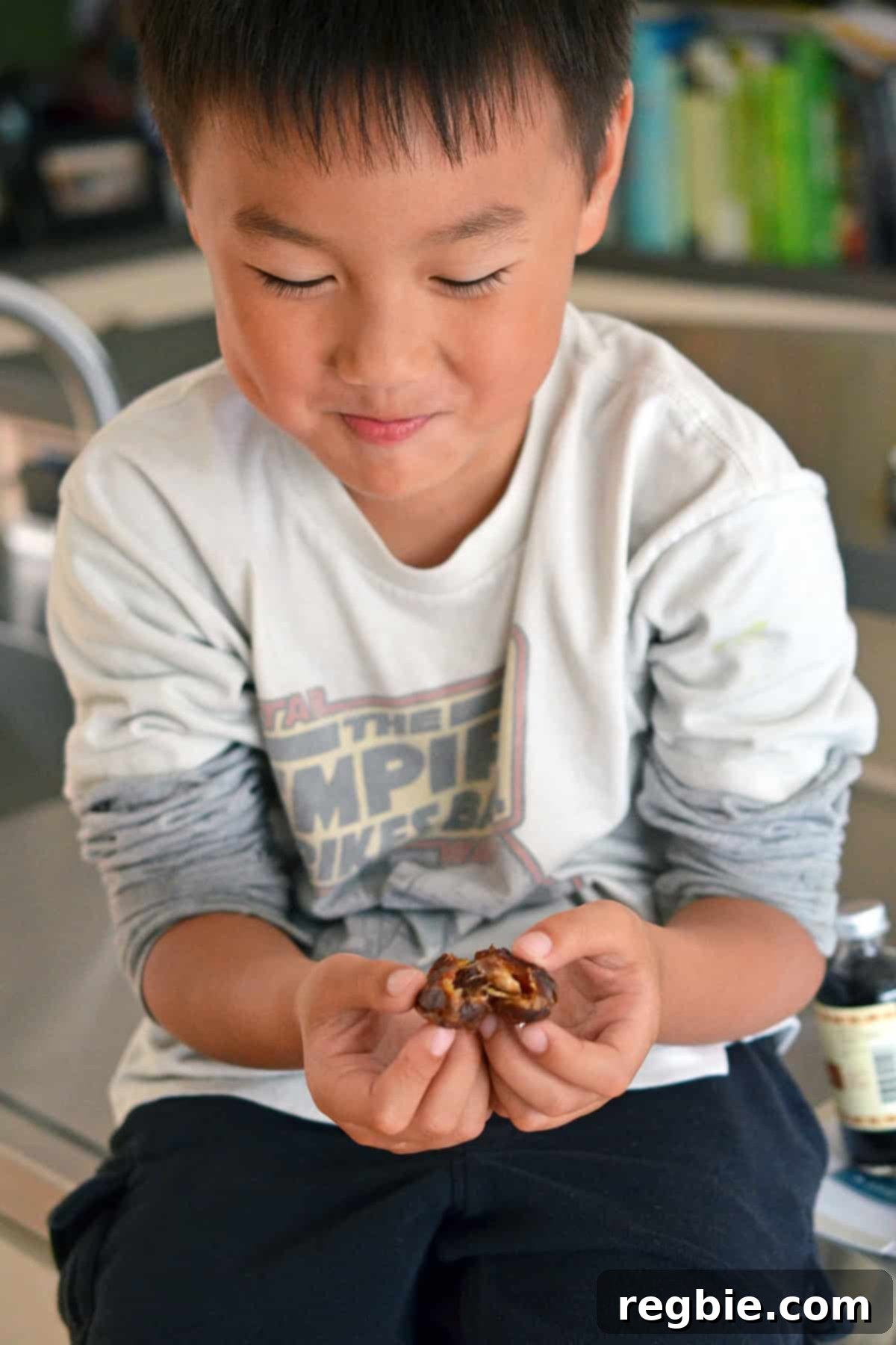 A smiling Asian boy sitting on a kitchen counter, carefully opening a Medjool date to remove the seed, assisting in the recipe preparation.