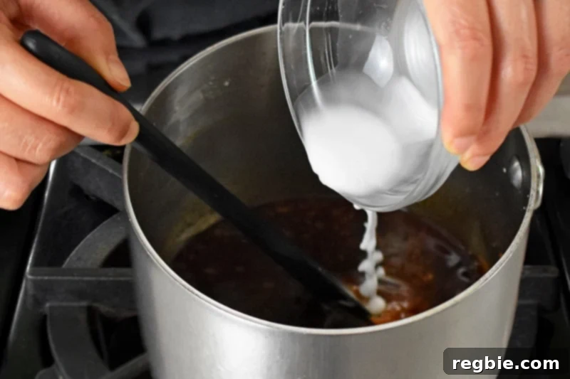 The precise action of pouring an arrowroot slurry into the Whole30 and paleo Asian sauce for sticky wings, illustrating the key moment of thickening.