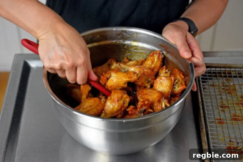 Someone expertly tossing crispy oven-baked chicken wings with a rich, maroon-colored spatula in a large mixing bowl, ensuring an even coating of the irresistible sticky sauce.