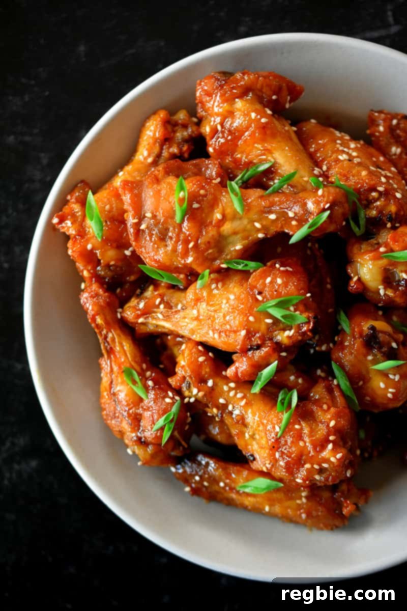 An inviting overhead shot of a large bowl brimming with perfectly coated Asian Sticky Wings, beautifully garnished with a generous sprinkle of toasted sesame seeds and vibrant sliced scallions, ready to be devoured.