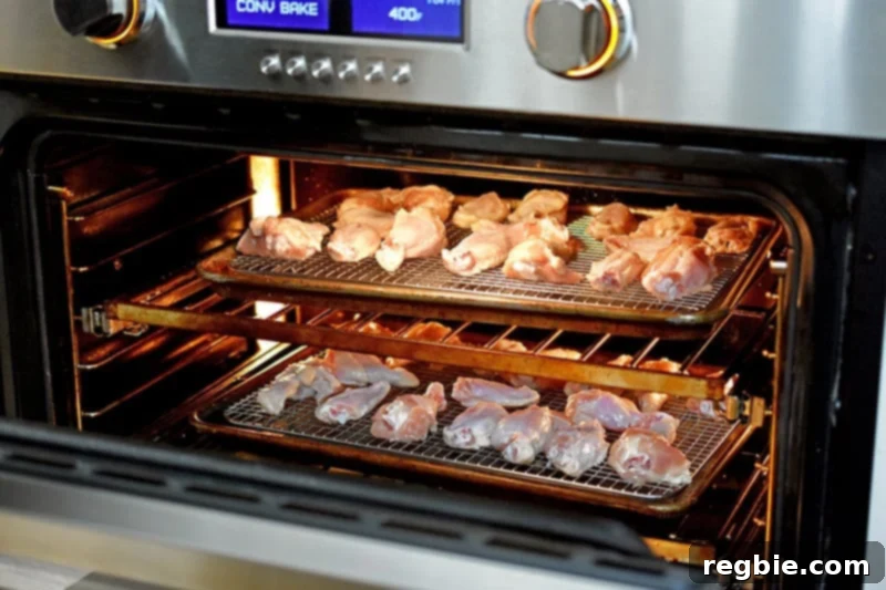 An open oven reveals two trays of chicken wings, strategically positioned on different racks, midway through their initial baking cycle, gently beginning to brown.