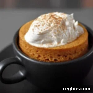 A black coffee mug filled with paleo and gluten-free pumpkin mug cake cooked in the microwave.The cake is topped with whipped coconut cream and dusted with pumpkin spice blend.