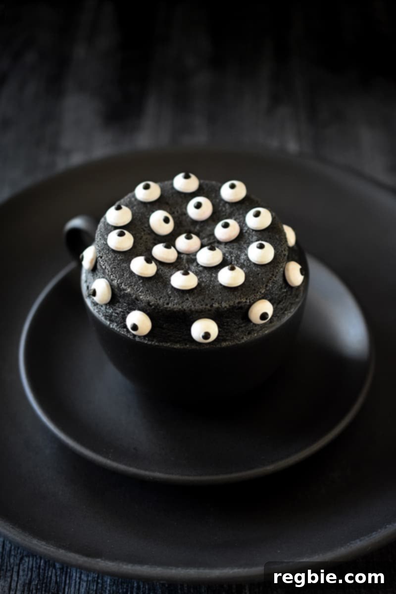 Paleo Black Sesame Mug Cake Indulgence 12 A black mug on two black plates is filled with a black sesame cake, richly topped with candy eyeballs, creating a fun and spooky dessert presentation.