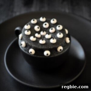 A black mug on two black plates is filled with a black sesame cake topped with candy eyeballs.
