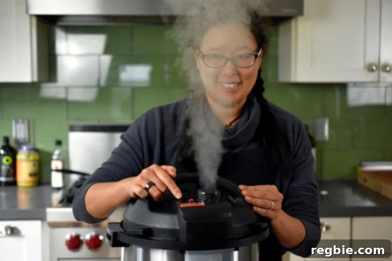 A dark haired smiling woman is manually releasing the pressure on an Instant Pot and the steam is right in front of her face.