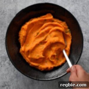 An overhead shot of a hand scooping vegan and paleo Instant Pot Mashed Sweet Potatoes from a black bowl. The vibrant orange mash is smooth and inviting.