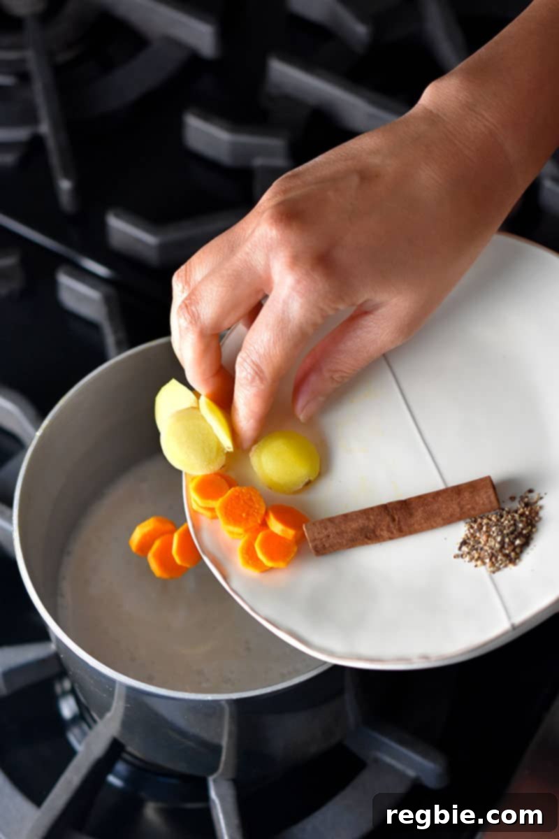 Paleo Dairy-Free Golden Turmeric Elixir 3 A hand adding turmeric slices, ginger slices, a cinnamon stick, and ground pepper to a saucepan filled with coconut milk to make golden milk.