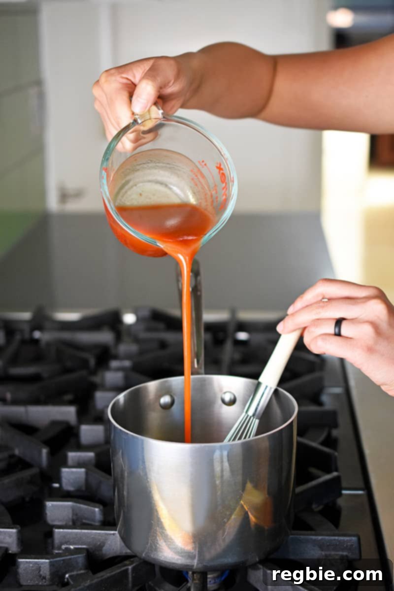 A hand is pouring bright red cayenne pepper sauce from a liquid measuring cup into a small saucepan, joining other ingredients to create the flavorful buffalo cauliflower sauce.