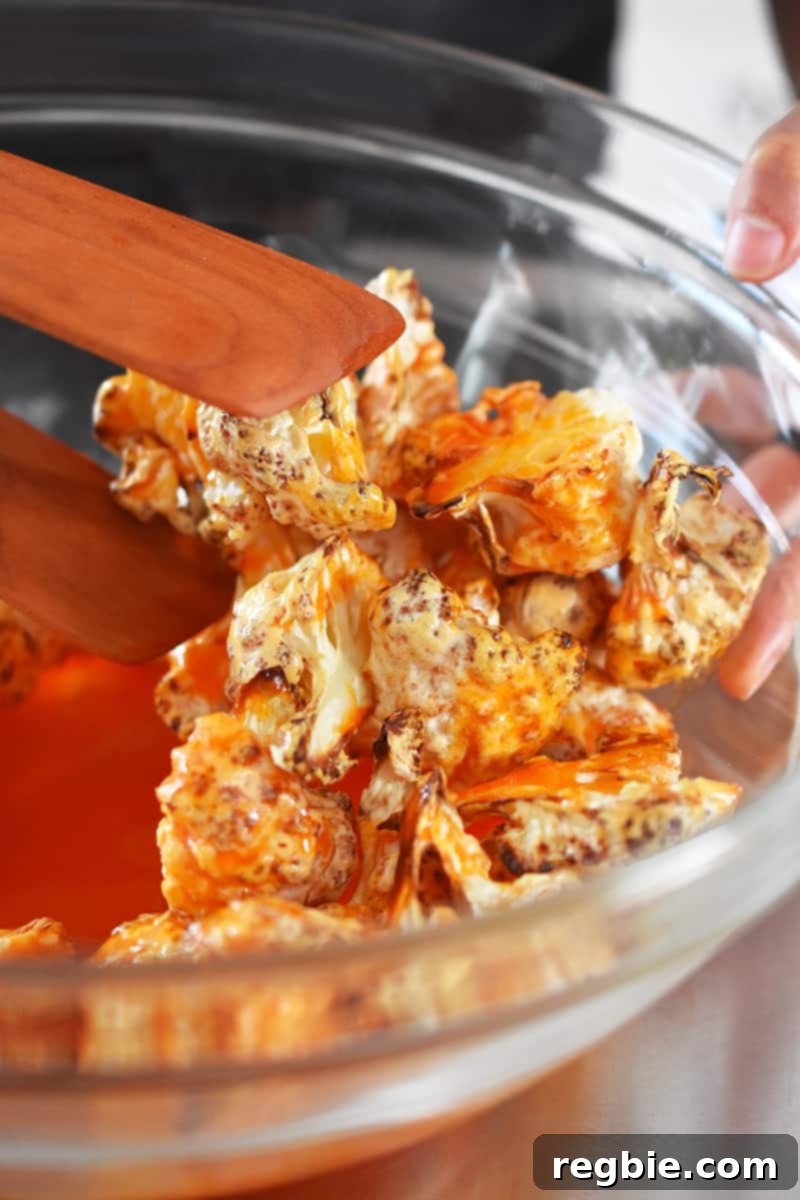 Crispy cauliflower florets being enthusiastically tossed with rich, red buffalo sauce in a clear glass bowl, ensuring every piece is perfectly coated and vibrant.