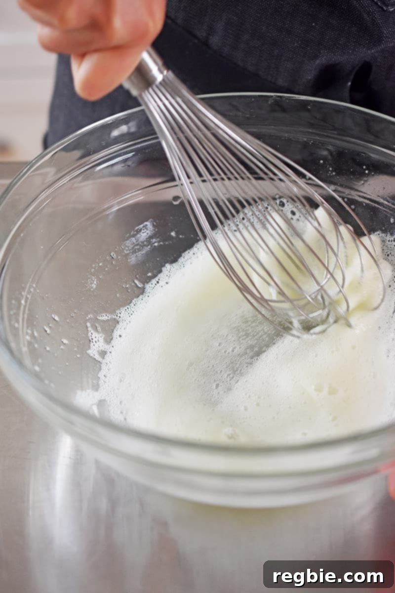 A whisk vigorously whipping egg whites in a clear glass bowl until they are frothy and bubbly, forming a light and airy base for the batter.
