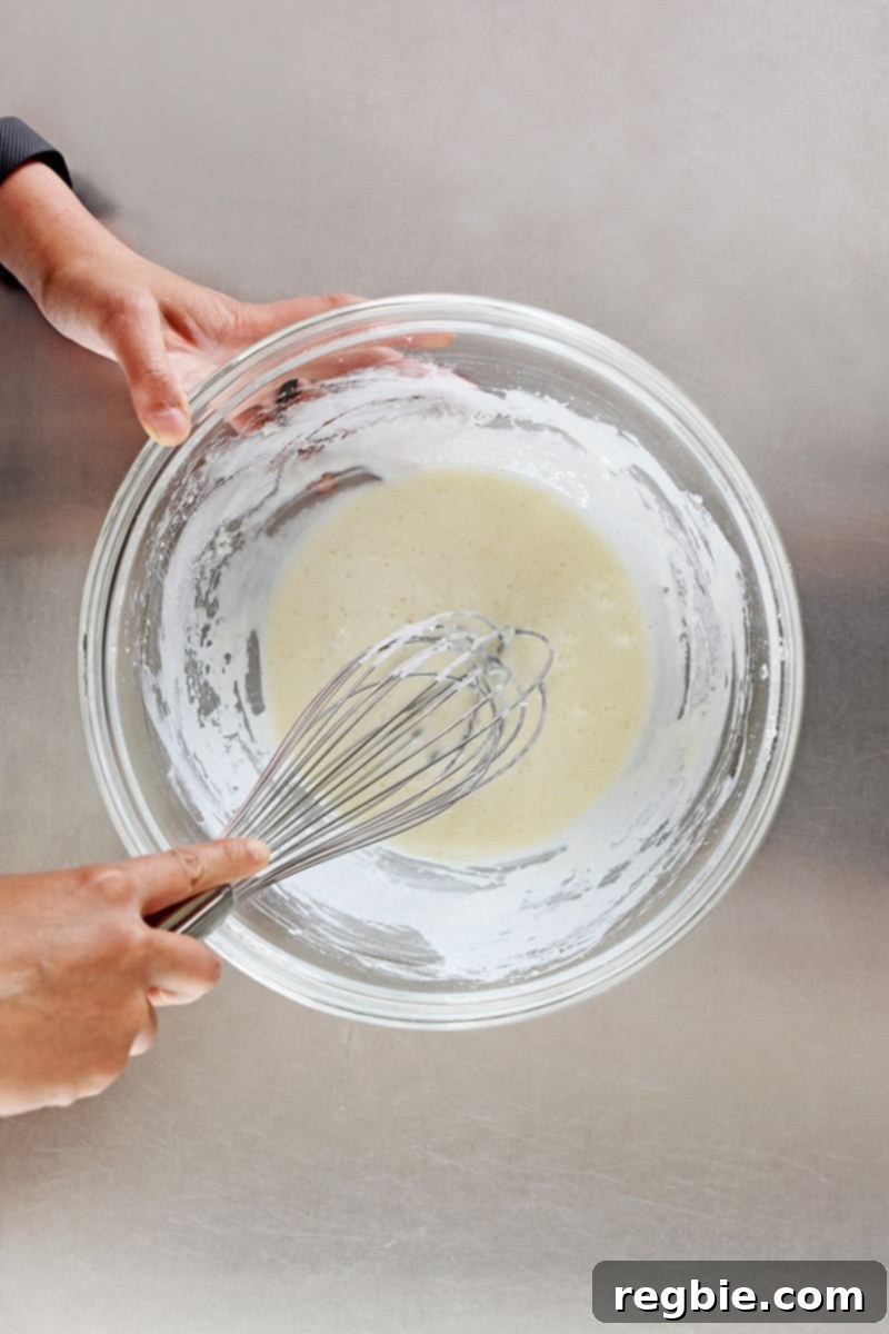 An overhead shot of a hand whisking the smooth, sticky batter for Nom Nom Paleo's Baked Buffalo Cauliflower in a clear glass bowl, ensuring all dry ingredients are incorporated.