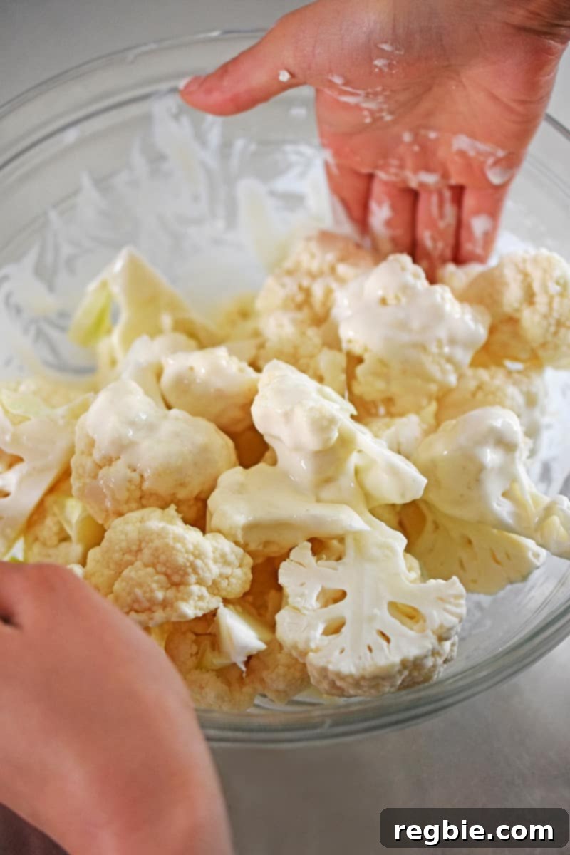Two hands are actively tossing cauliflower florets within the gluten-free, paleo, and Whole30 batter in a large bowl, ensuring an even and thorough coating on each piece.