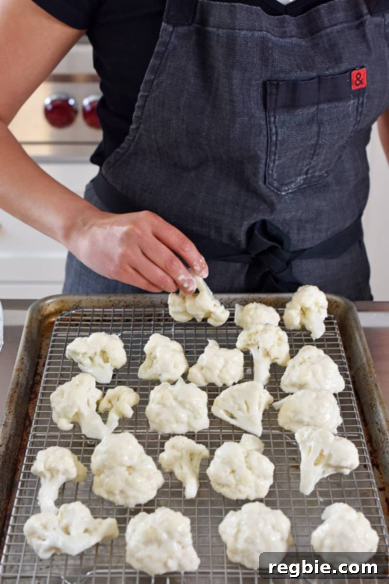 Someone in a gray apron carefully placing battered cauliflower florets onto a wire rack set within a rimmed baking sheet, ensuring single layer placement for optimal baking.