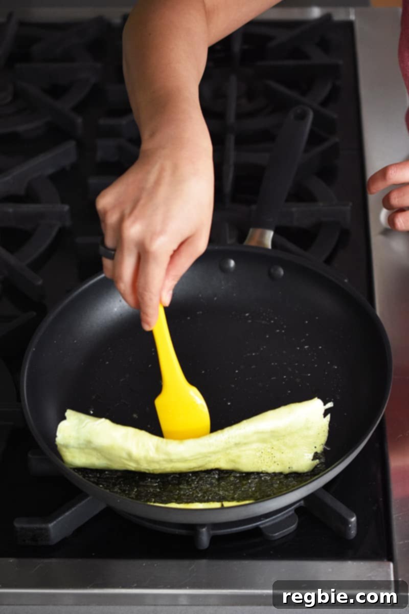 A hand is using a yellow silicone spatula flipping a Korean egg roll with roasted seaweed. The Korean egg roll is almost flipped all the way at the end.