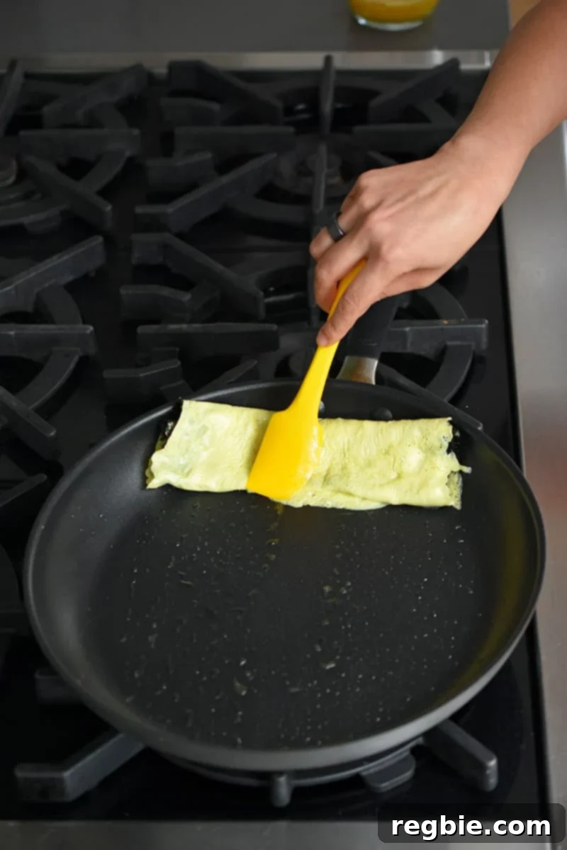 A hand holding a yellow silicone spatula is pushing a rolled egg omelet with seaweed to the edge of a large non-stick skillet.