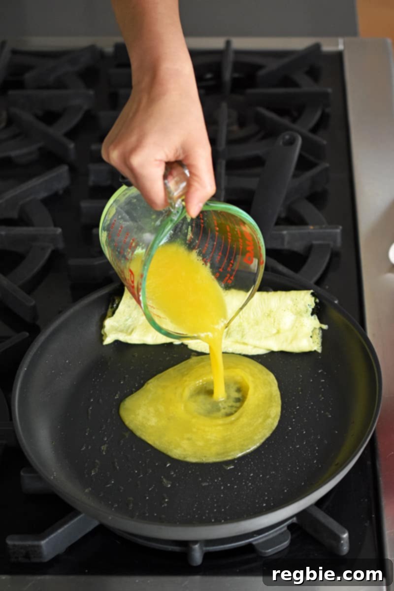 A hand pours whisked eggs from a measuring cup into a non-stick skillet that has a cooked egg roll omelet on the edge.