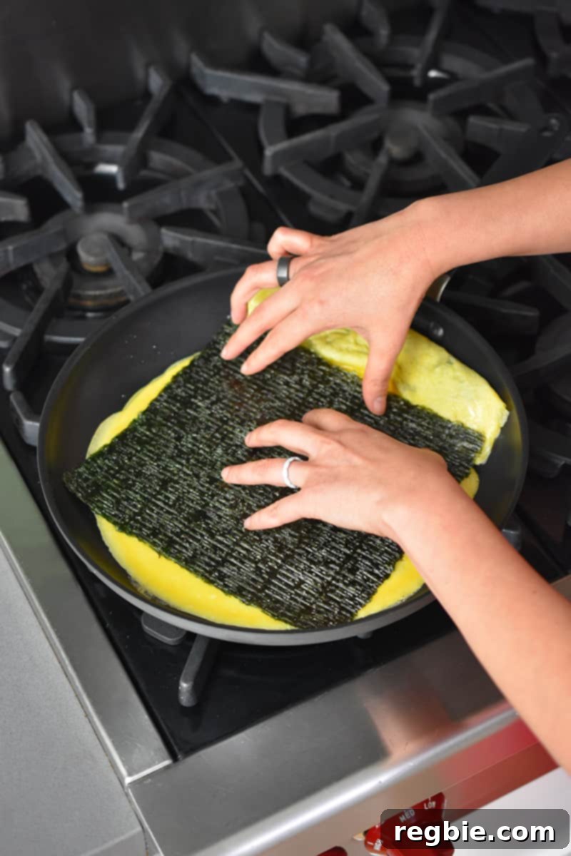 Two hands are placing a rectangular roasted seaweed in a skillet filled with a thin egg omelet.