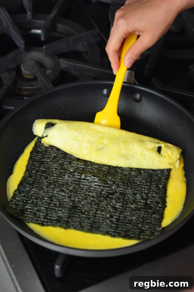A hand is using a yellow silicone spatula to start flipping a Korean egg roll.