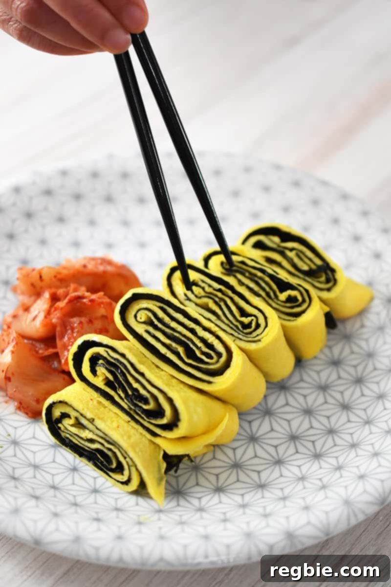 A pair of chopsticks is grabbing a slice of Korean egg roll with seaweed on a white and gray plate. There is a small pile of kimchi next to it.