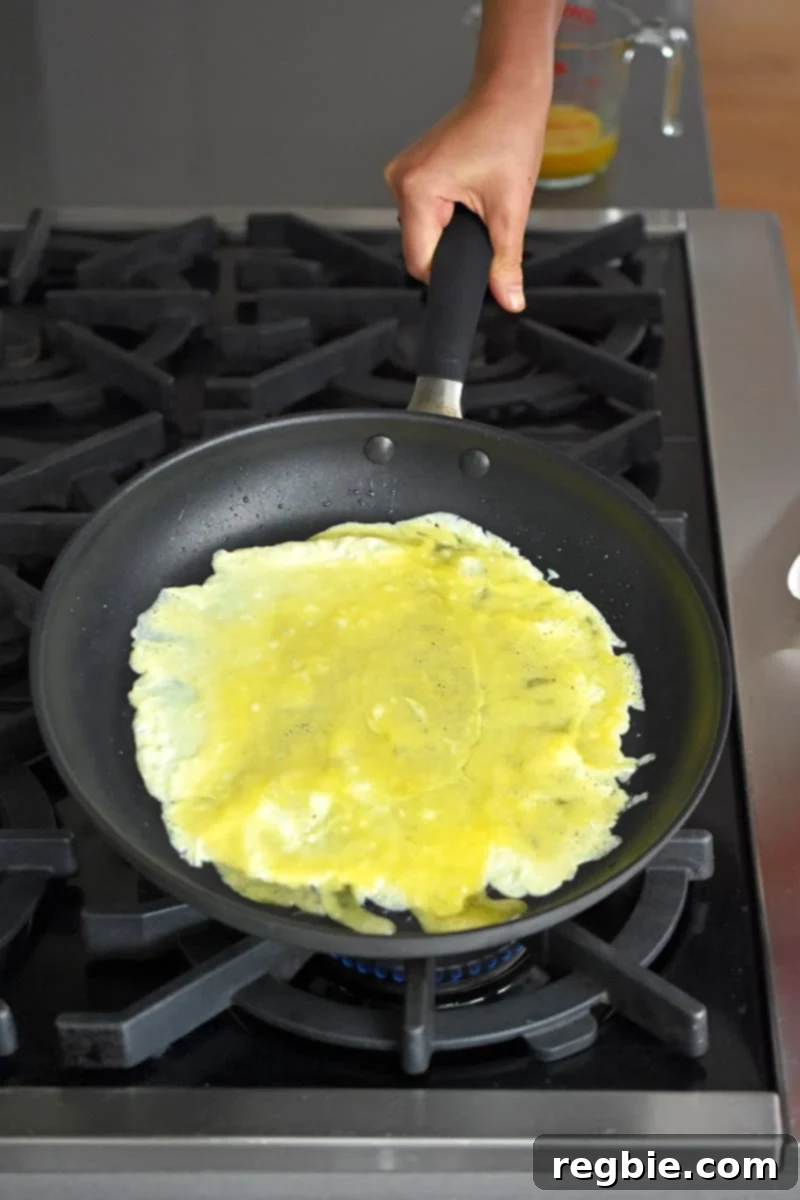 A hand is tipping a large non-stick skillet to make a thin layer of egg omelet on the bottom of the skillet.