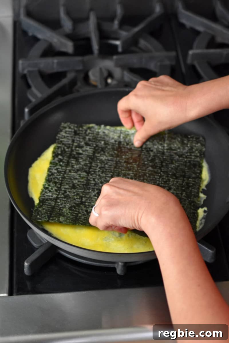 Two hands placing a rectangular piece of roasted seaweed on a thin layer of cooked egg in a non-stick skillet.