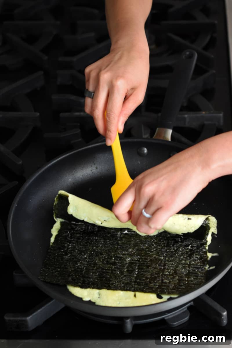 Two hands are using a yellow silicone spatula to fold over the edge of an egg omelet topped with seaweed.