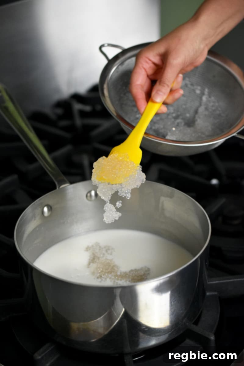 Tropical Mango Coconut Tapioca 14 A yellow spatula is spooning cooked small tapioca pearls into a saucepan filled with sweetened coconut milk.