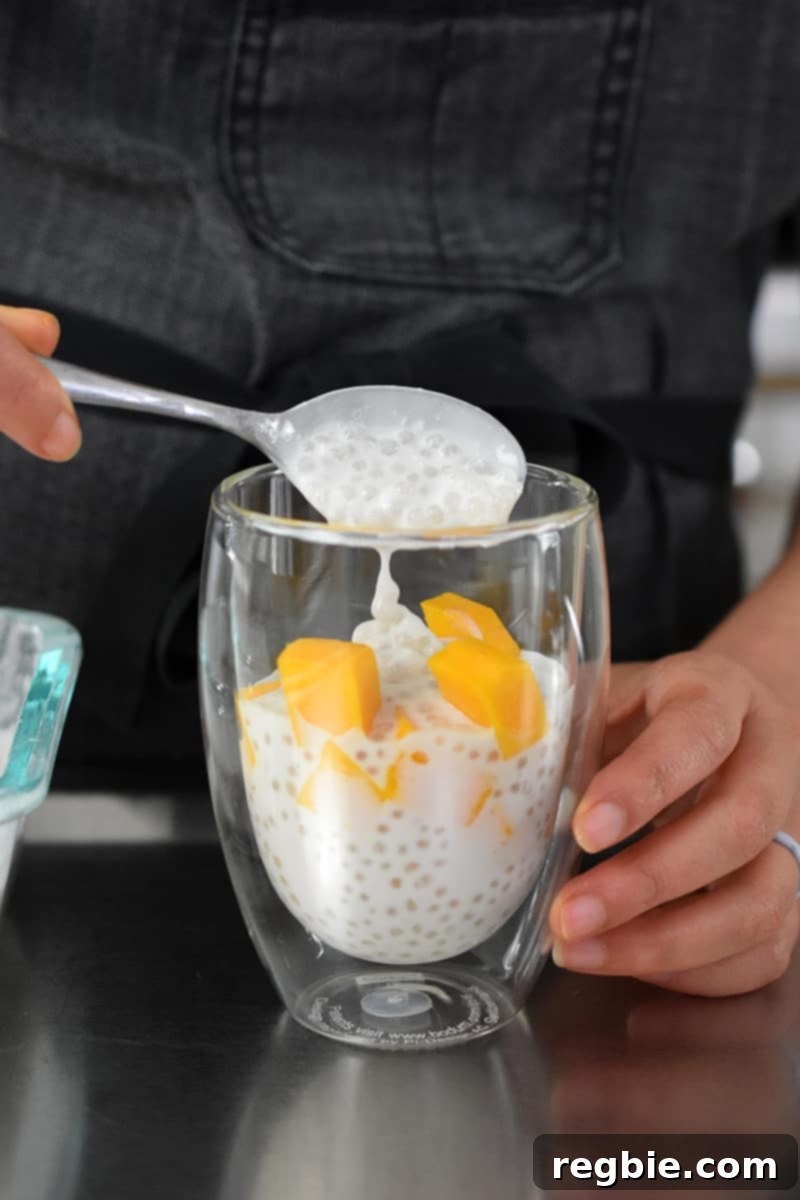 Tropical Mango Coconut Tapioca 19 A front shot of someone layering coconut sago with diced mango in a clear glass cup.