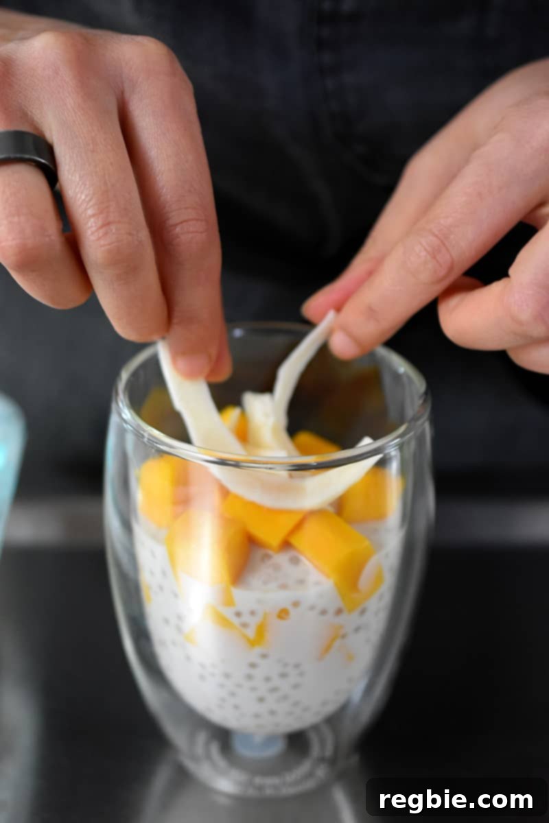 Tropical Mango Coconut Tapioca 4 Two hands adding toasted coconut on top of clear glass filled with mango sago, a coconut tapioca pudding with diced mango
