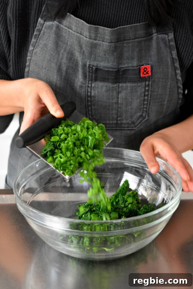 Garden Colcannon 12 A person in a gray apron is transferring chopped kale and scallions with a bench scraper into a large glass bowl.
