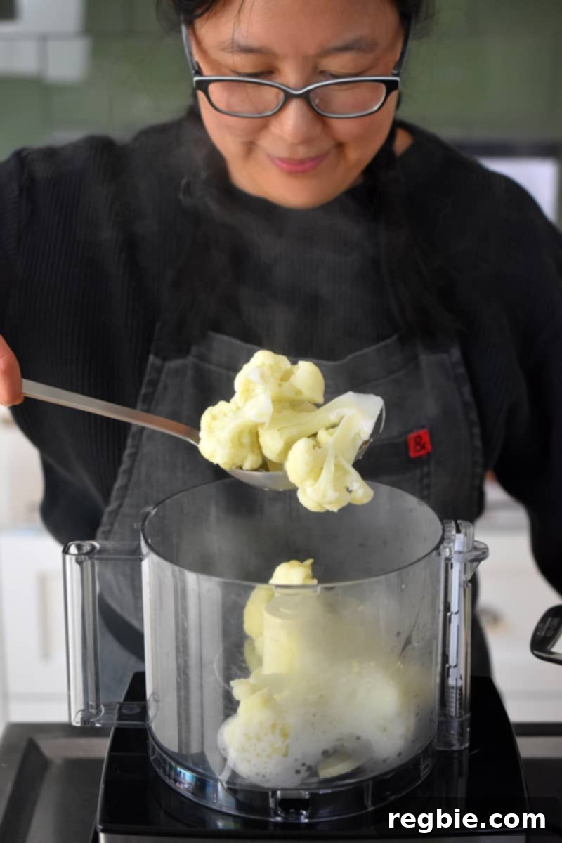 Garden Colcannon 13 A smiling Asian woman is putting cooked cauliflower florets into an open food processor.