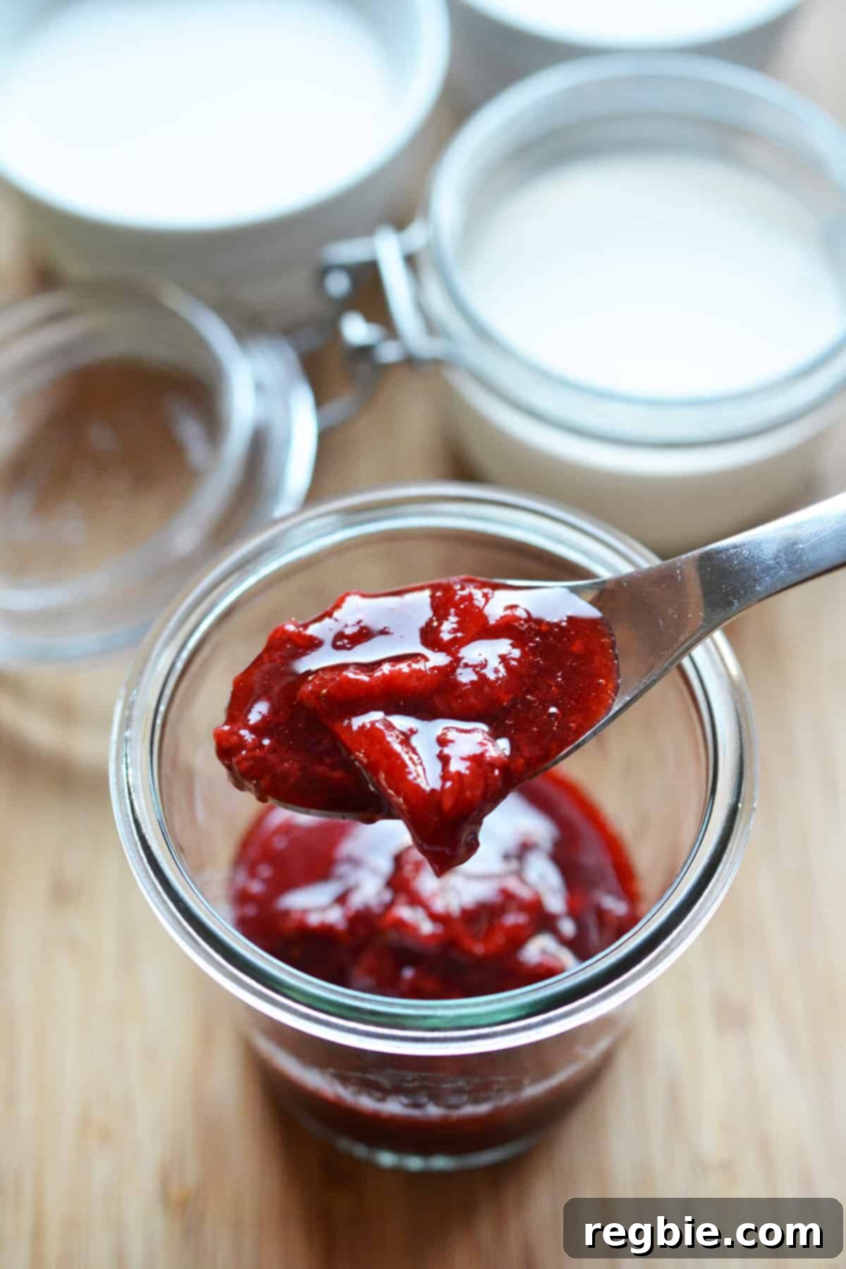 A spoonful of strawberry compote from a glass jar filled with it.