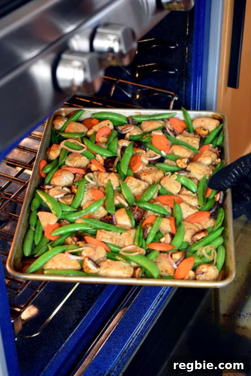 One-Pan Wonder Chicken Stir-Fry 12 A gloved hand is rotating a sheet pan chicken and vegetable dinner at the midway point while it is baking.