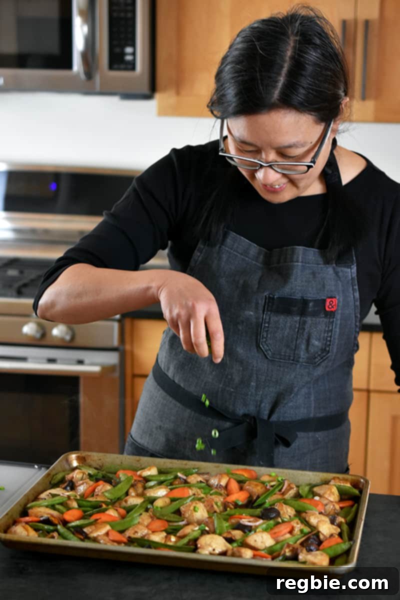 One-Pan Wonder Chicken Stir-Fry 14 An Asian woman is sprinkling sliced scallions on a sheet pan filled with stir-fry chicken and vegetables.