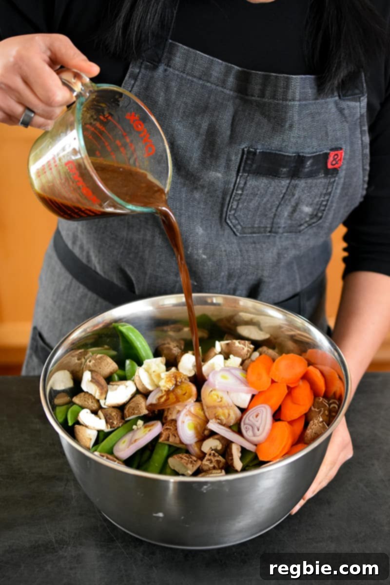 One-Pan Wonder Chicken Stir-Fry 3 A person in a gray apron is pouring a brown sauce into a large metal bowl filled with chopped carrots, sugar snap peas, shallots, shiitake mushrooms, and minced garlic.