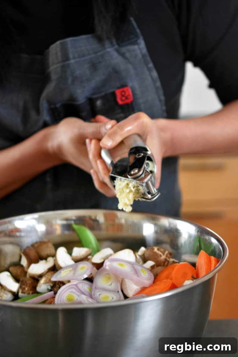 One-Pan Wonder Chicken Stir-Fry 7 Someone in a gray apron is squeezing a garlic press with two hands and adding the minced garlic to a big silver bowl filled with chopped vegetables