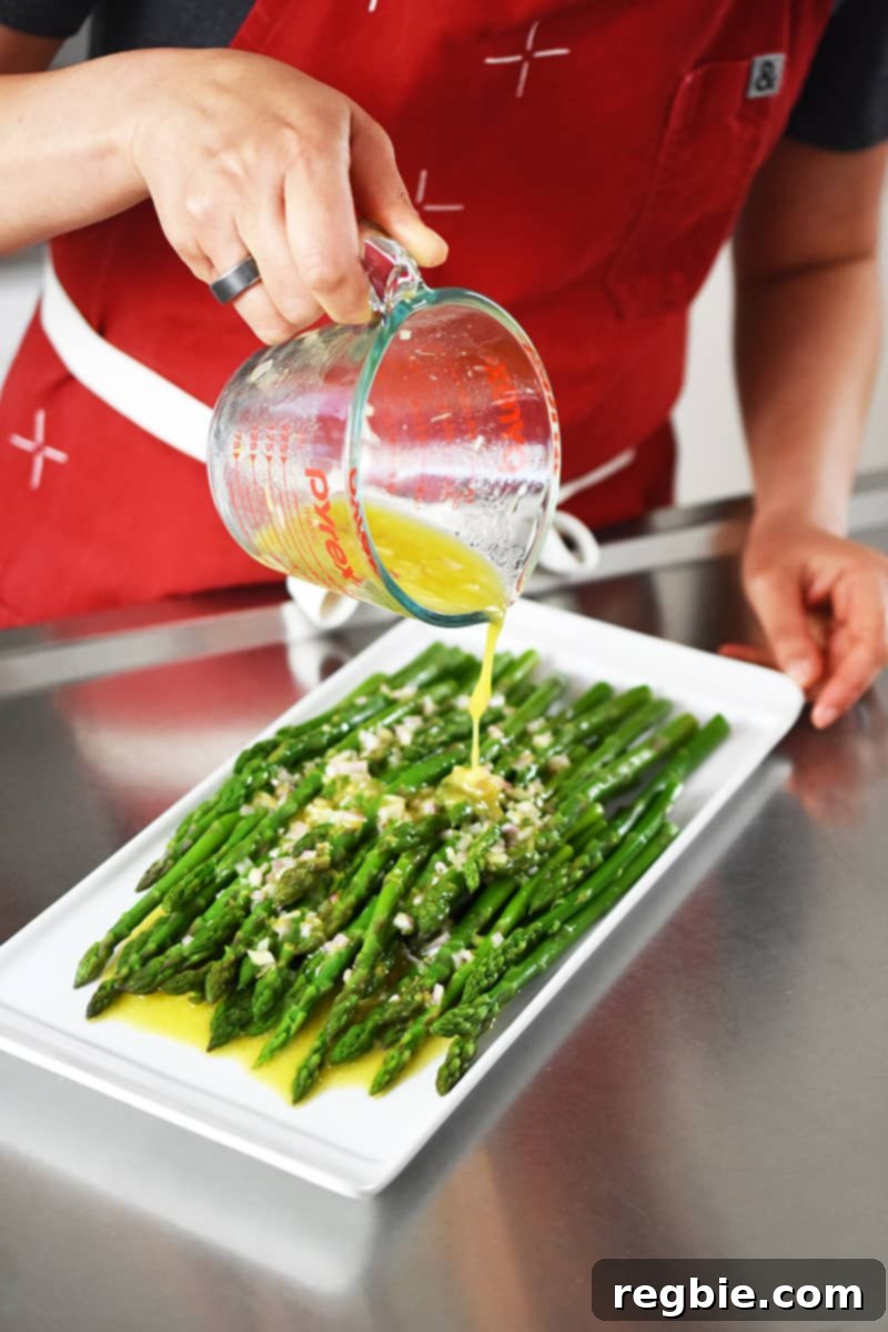 Whole30 Keto Paleo Asparagus Mimosa 16 Someone in a red apron is pouring a lemon and shallot vinaigrette on top of cooked asparagus on a white rectangular plate.