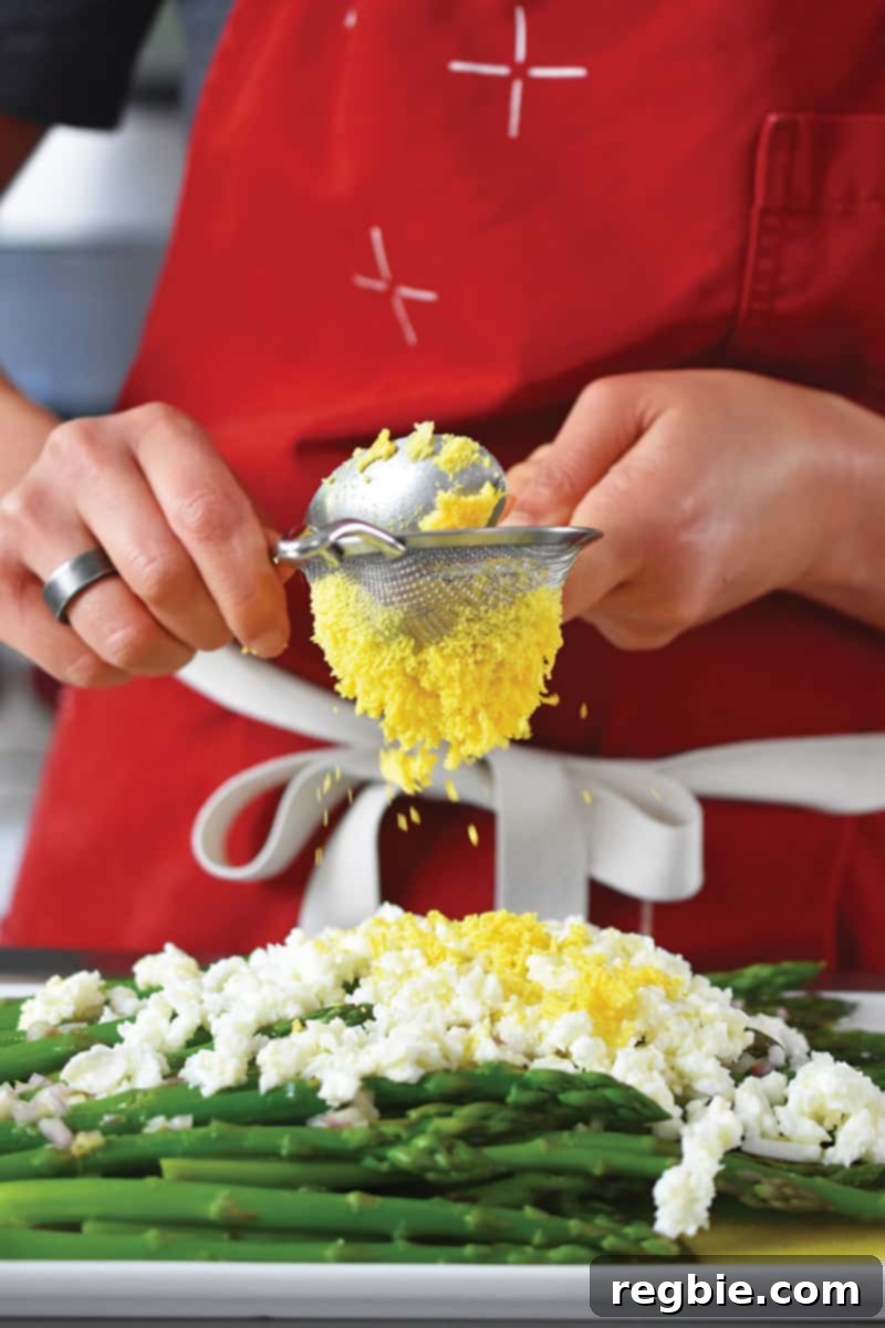 Whole30 Keto Paleo Asparagus Mimosa 18 Someone in a red apron is pushing cooked egg yolks though a small mesh sieve onto a plate of asparagus topped with egg whites.