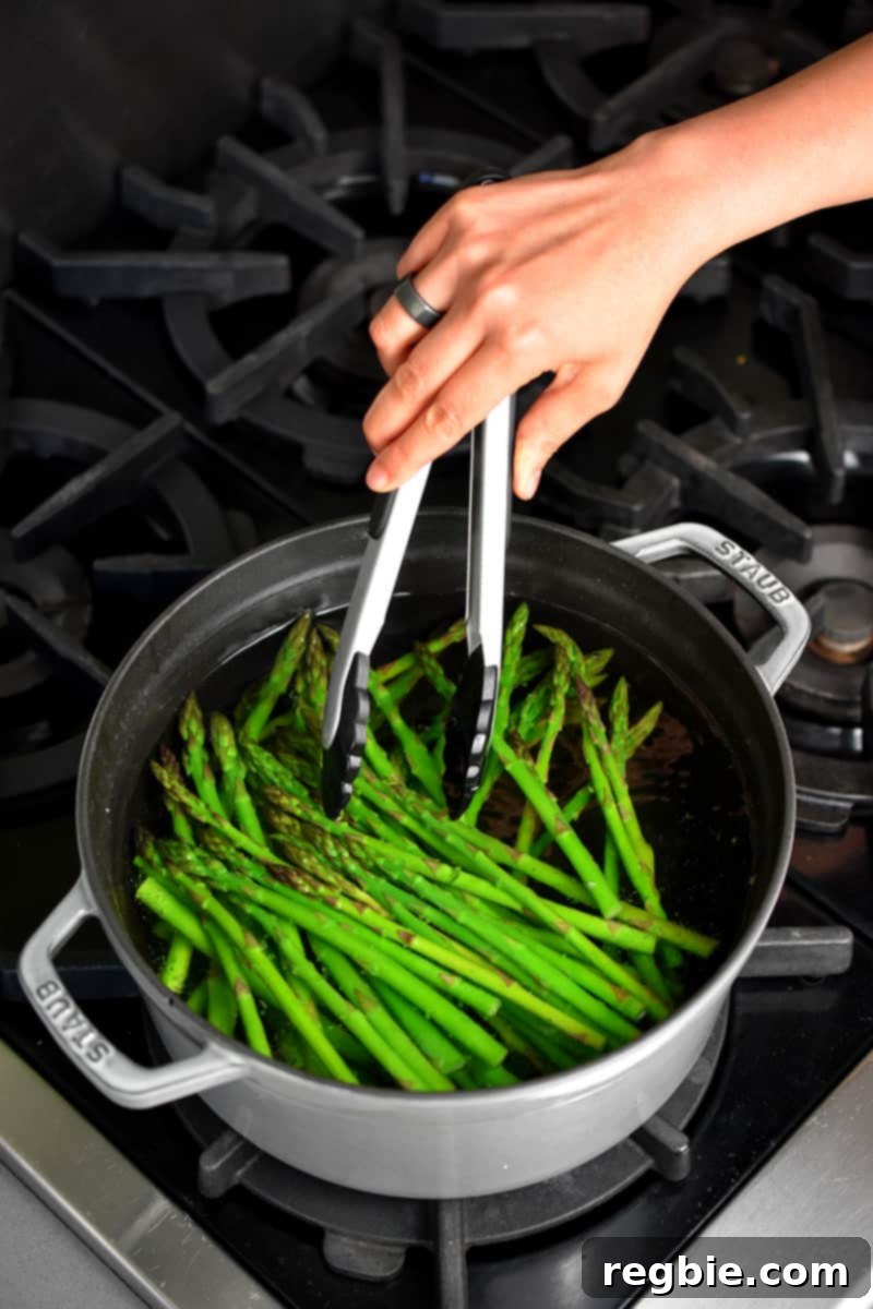 Whole30 Keto Paleo Asparagus Mimosa 10 An overhead shot of a gray pot filled with boiling water and bright green asparagus. A hand with a pair of tongs is mixing them around.