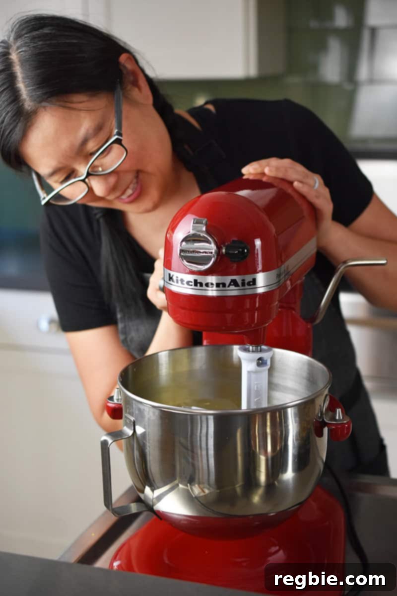 A smiling Asian woman is actively blending the light pink strawberry muffin batter in a vibrant red KitchenAid stand mixer, ensuring all ingredients are well incorporated.
