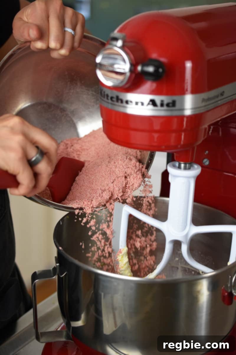 The process of carefully pouring the pink-colored dry ingredients for strawberry muffins into the bowl of a stand mixer, where the wet ingredients are already being blended.
