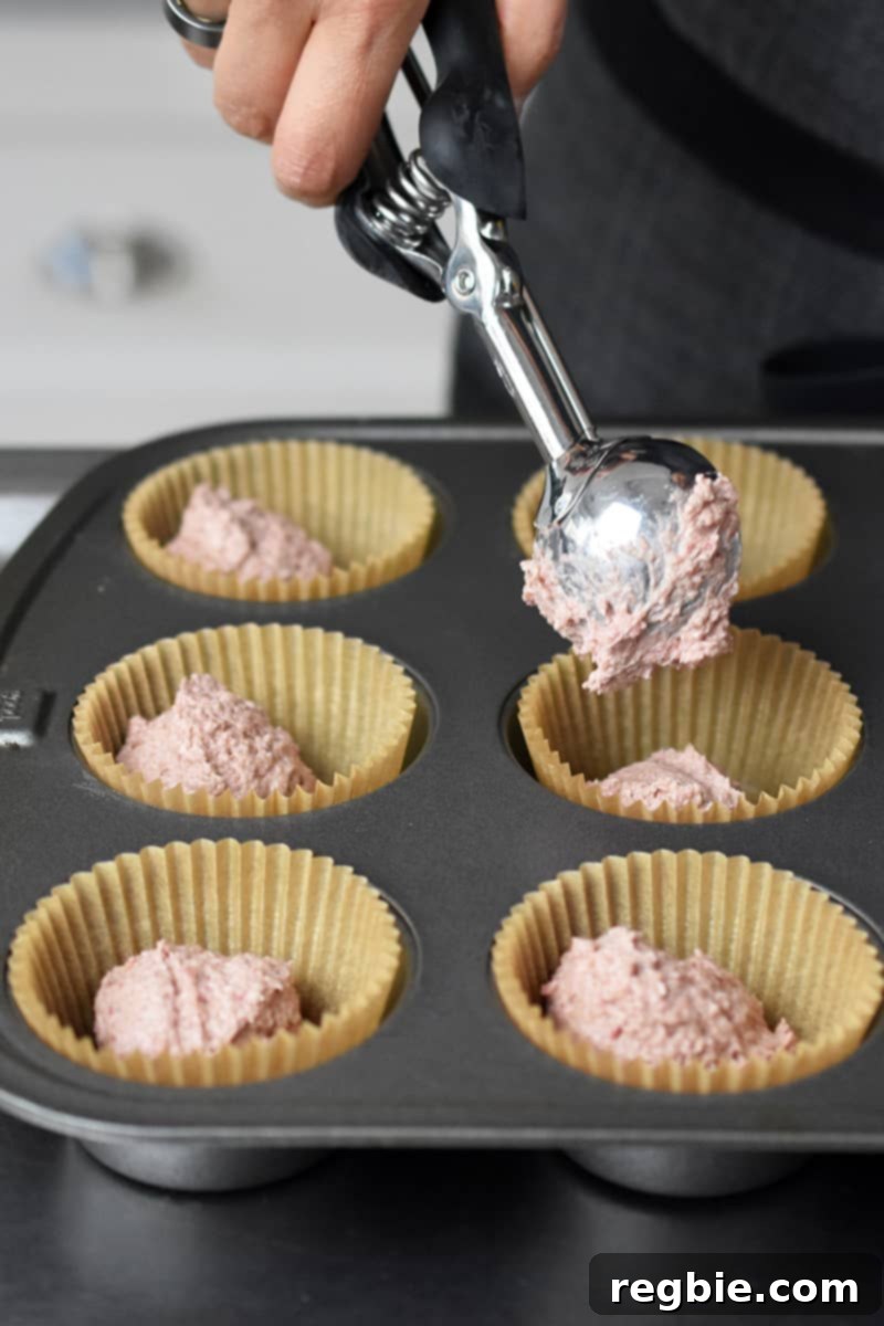 Someone carefully adds a small scoopful of the pink muffin batter to the bottom of a parchment-lined muffin compartment, creating a foundational layer.